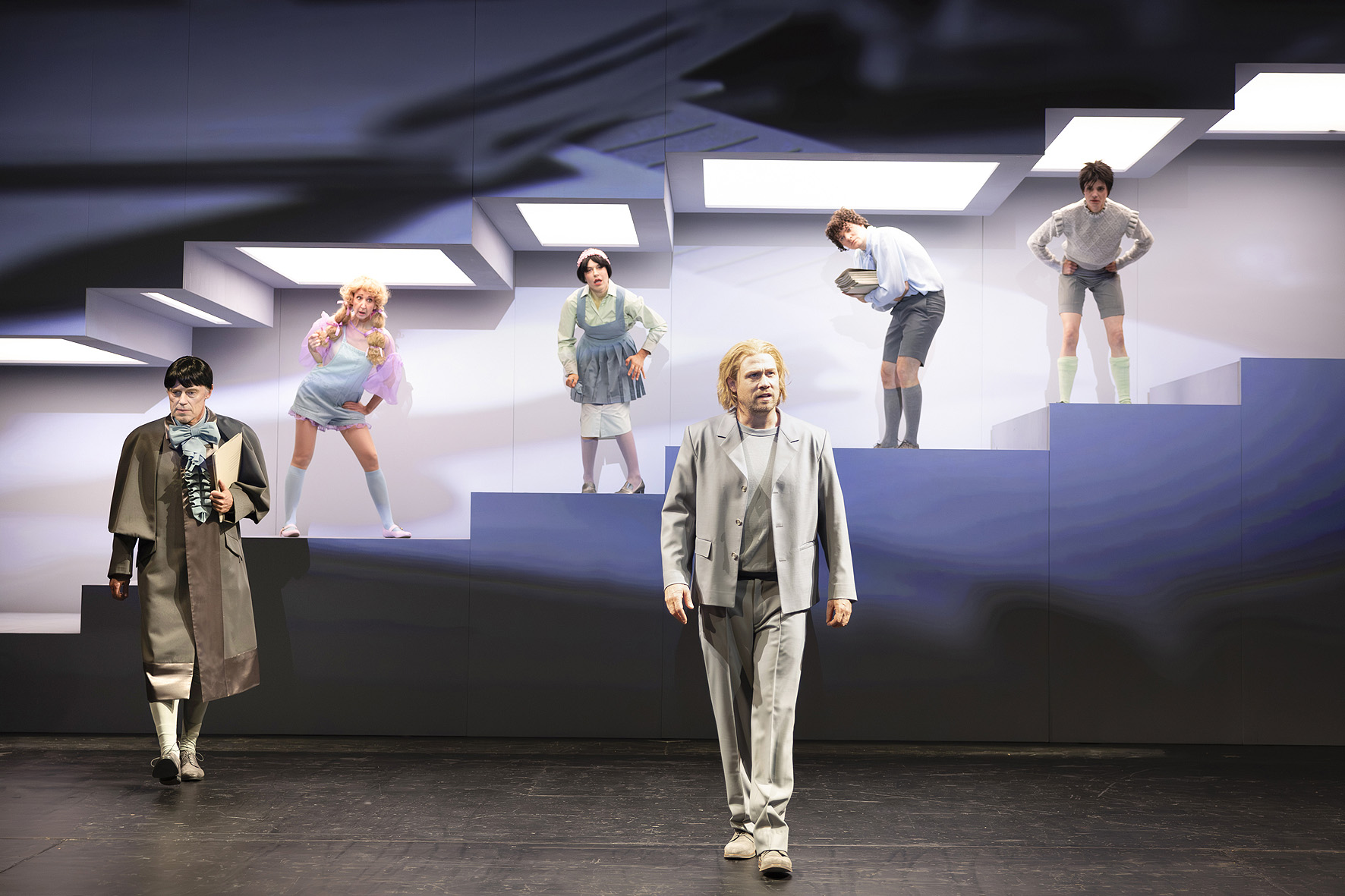 Bühnenszene aus „Der Prozess“ am Theater Lübeck: Figuren in stilisierten Kostümen stehen auf treppenartigen Ebenen vor einem hellen Bühnenbild.